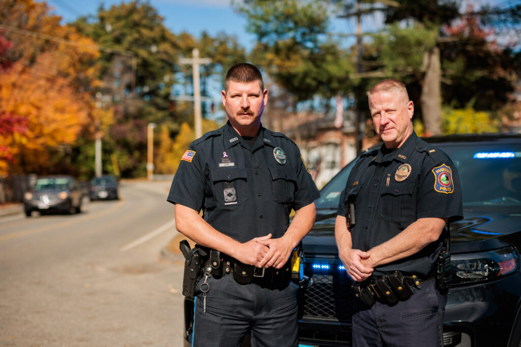 Two white male police officers stand in front of a police cruiser.