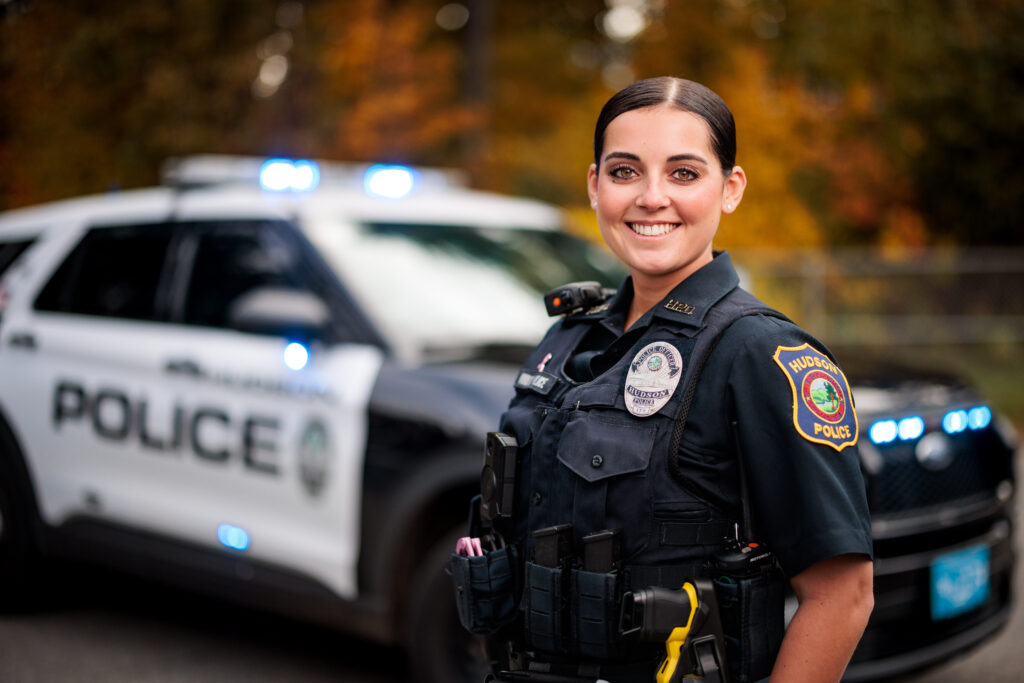 A white female police officer stands in front of a police cruiser.