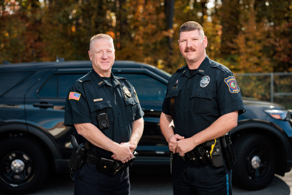 Two white male police officers standing in front of a police cruiser.