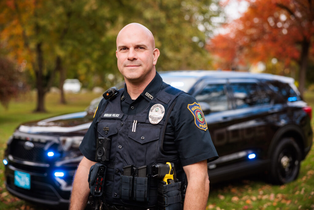 A white male police officer stands in front of a police cruiser.