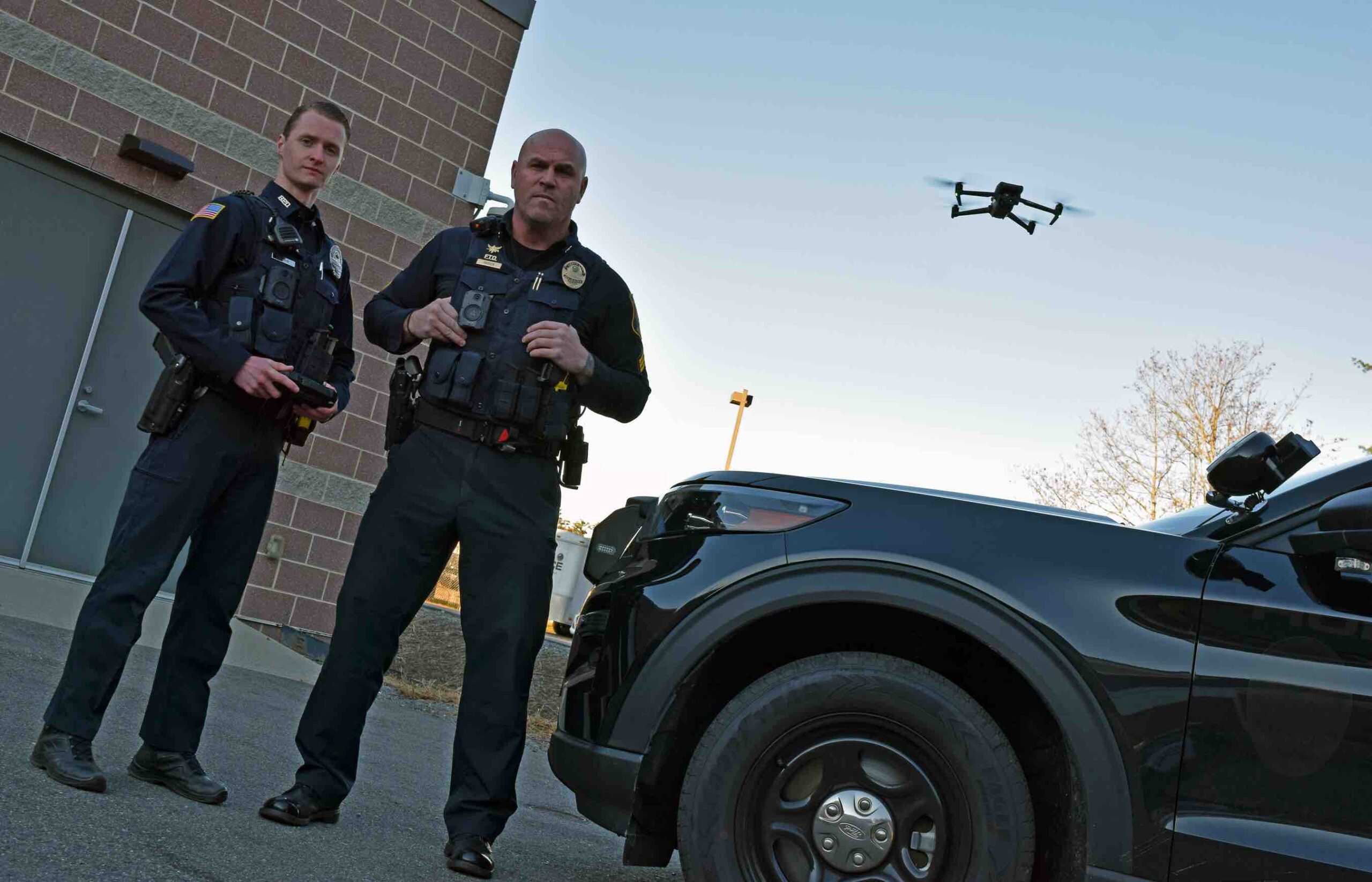 Two Hudson Police Officers stand in front of a police car while a drone hovers behind them in the air.