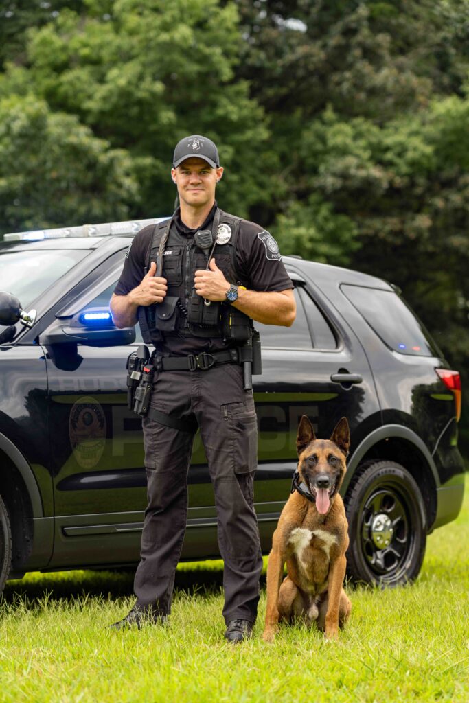 A male police officer stands in front of a cruiser with a Hudson K9.