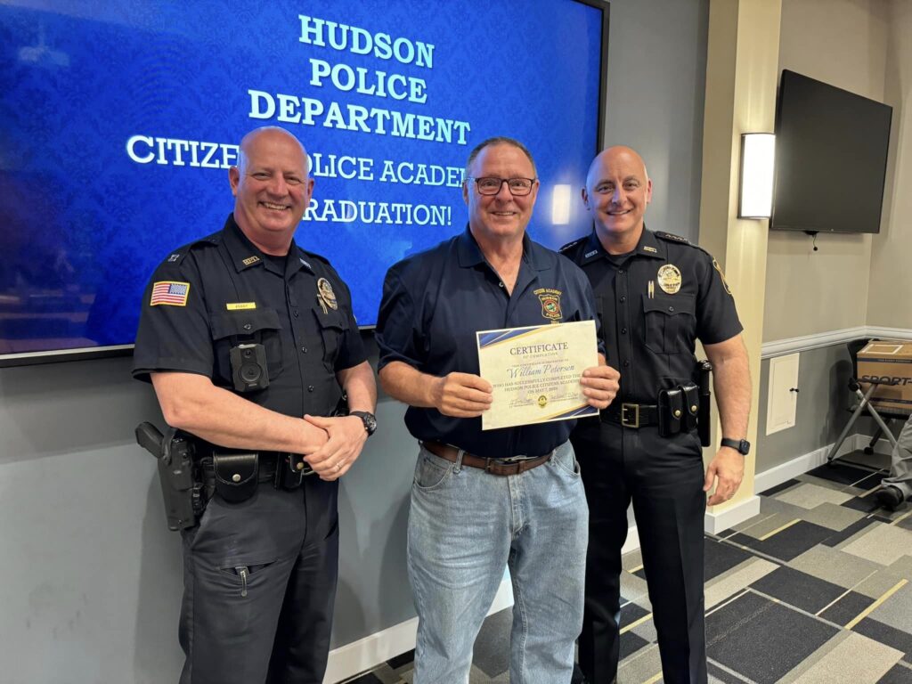 Citizens Police Academy participant holds a certificate while posing for a photo with two Hudson Police officers.